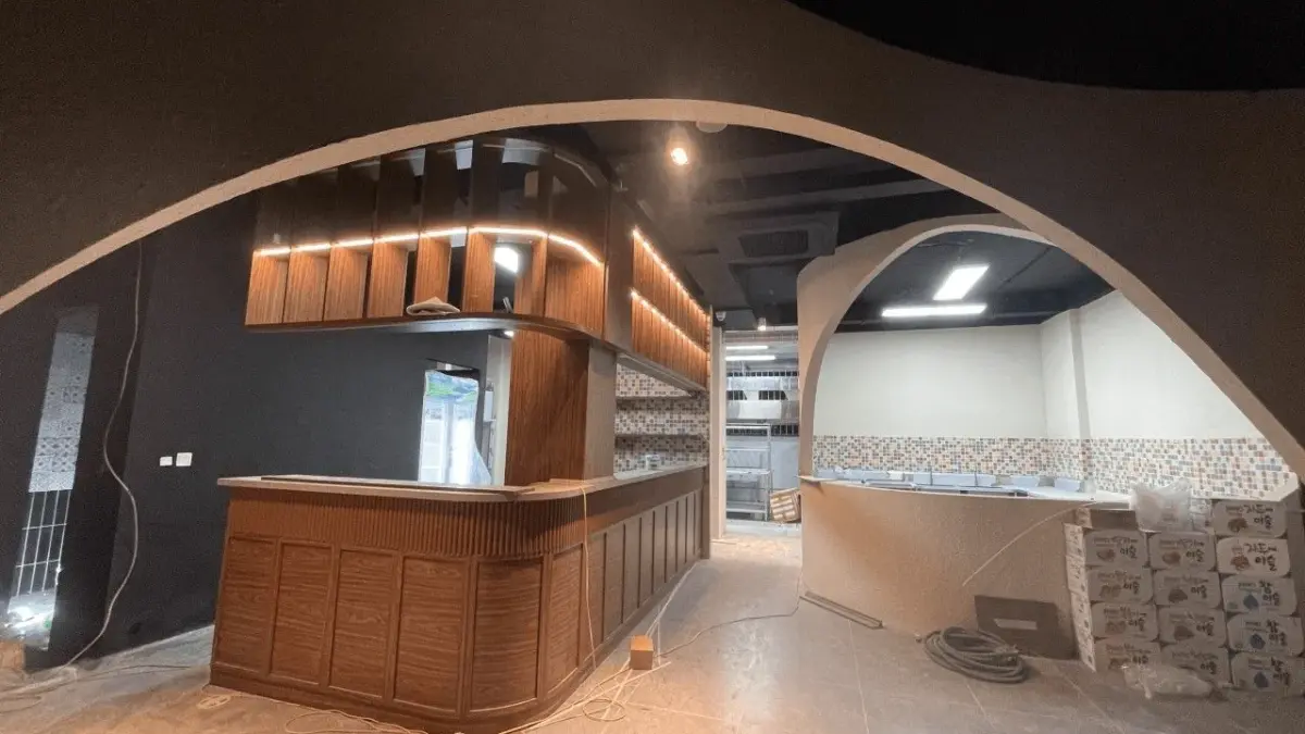 Ảnh thi công thực tế – quầy bar sushi gỗ óc chó đang hoàn thiện, hệ vòm cuốn thạch cao tạo hình hữu cơ đặc trưng, khu bếp ốp gạch mosaic phía sau