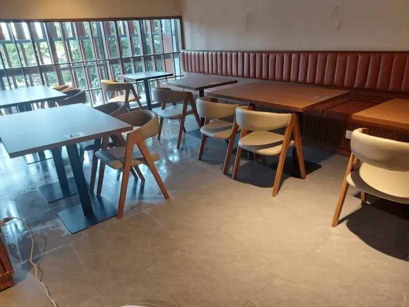 Ảnh thực tế hoàn thiện – khu bàn ăn với ghế băng sofa da nâu dọc tường, ghế gỗ chân vát, cửa kính lớn đón sáng và cây xanh bên ngoài